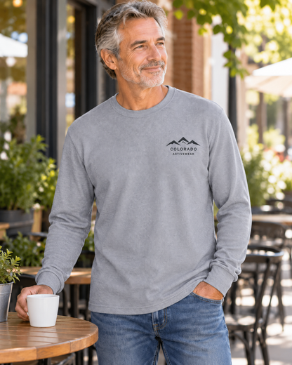 Illustrative lifestyle mockup: man wearing the Heather Gray Colorado Mountain Waffle Long Sleeve at an outdoor cafe, showing its small dark left-chest mountain logo.