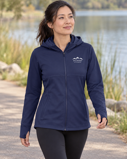 Illustrative lifestyle mockup: woman wearing the True Navy Women’s Mountain Hooded Softshell Jacket on a lakeside path, showing its small white left-chest mountain logo.