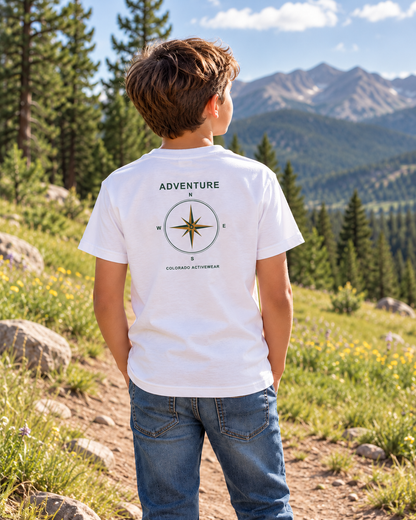 Illustrative lifestyle mockup: child wearing the White Colorado Adventure Compass Youth Tee on a meadow path, showing the compact green and gold compass back graphic.