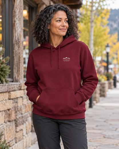 Illustrative lifestyle view of a woman wearing the Burgundy Alpine Social Club Fleece Hoodie on a mountain-town sidewalk, showing the small white Colorado Activewear chest mark.