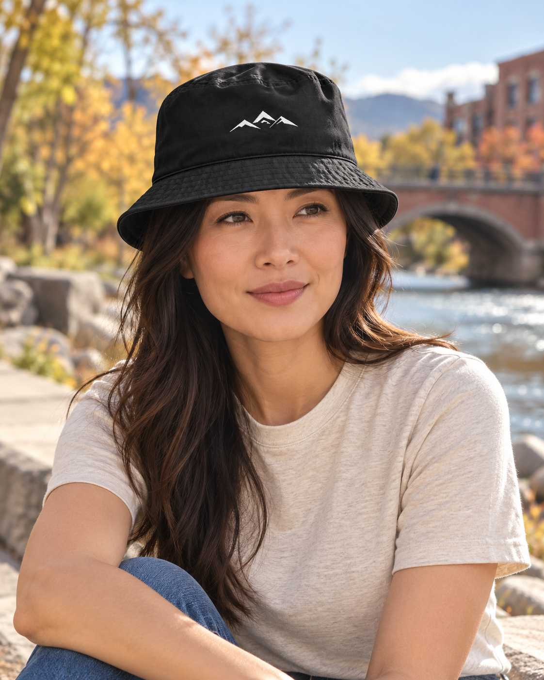 Illustrative lifestyle view of a woman wearing the Black Colorado Mountain Embroidered Bucket Hat beside a riverside path.
