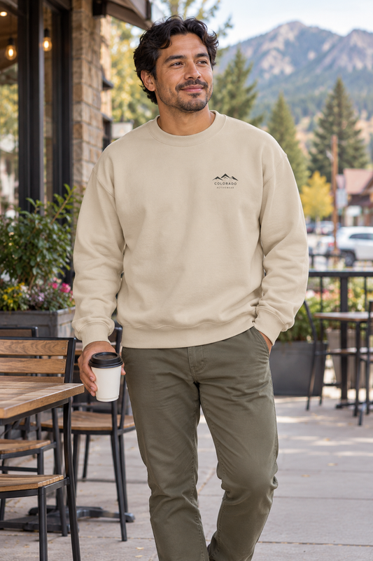 Illustrative lifestyle mockup: man wearing the Sand Colorado Mountain Logo Crewneck at a mountain-town cafe, showing its small dark left-chest logo.