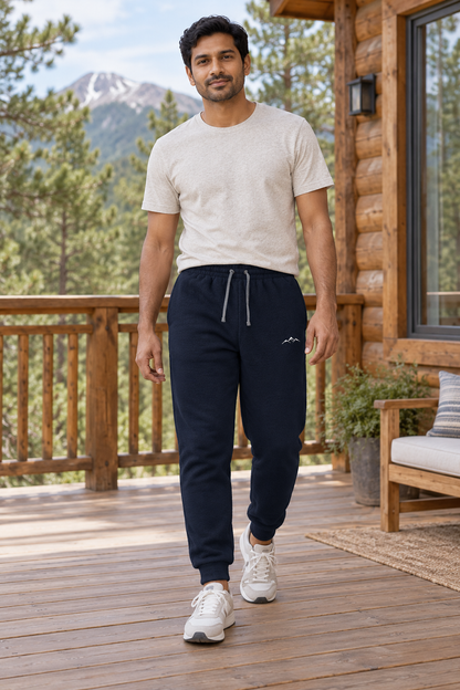 Illustrative lifestyle mockup: man wearing the J. Navy Colorado Mountain Embroidered Joggers on a cabin deck, showing the small white mountain thigh logo, drawcord and cuffed ankles.