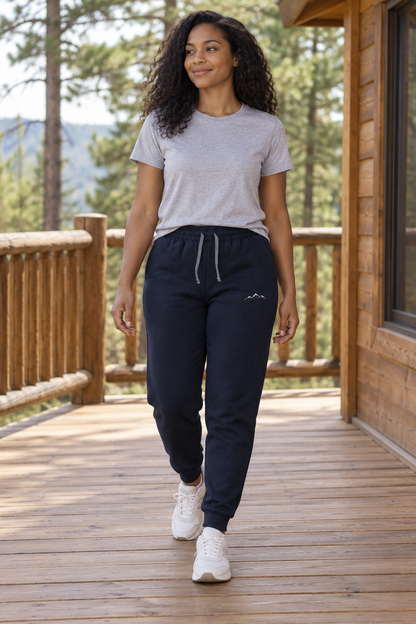 Illustrative lifestyle mockup: woman wearing the J. Navy Colorado Mountain Embroidered Joggers on a cabin deck, showing the small white mountain thigh logo, drawcord and cuffed ankles.