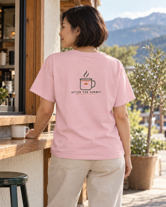 Blossom After the Summit Camp Mug Tee on a woman at an outdoor cafe, showing the compact mug-and-steam back graphic; illustrative lifestyle mockup.