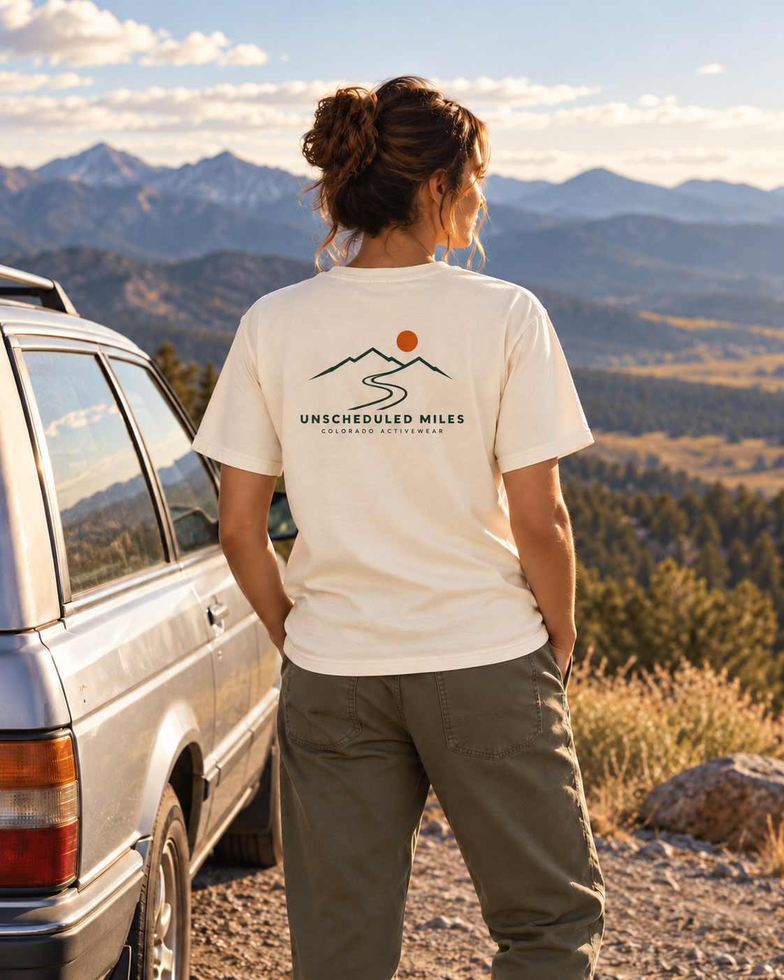 Illustrative lifestyle mockup: woman wearing the Ivory Unscheduled Miles Road Tee beside a station wagon at a mountain turnout, showing its compact back graphic.
