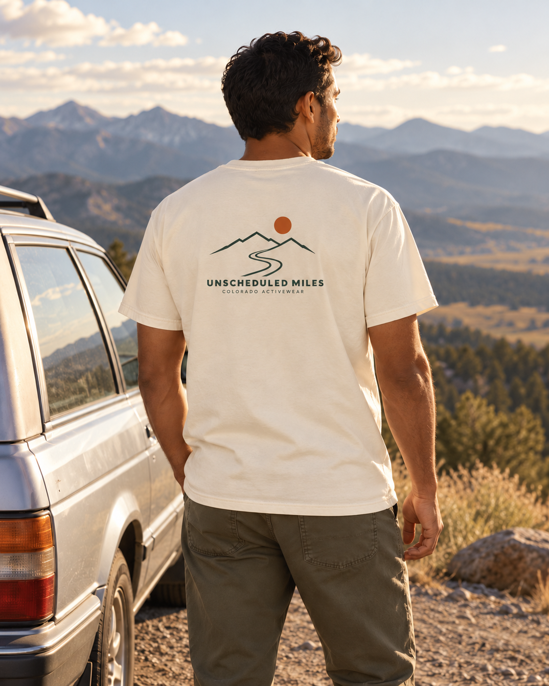 Illustrative lifestyle mockup: man wearing the Ivory Unscheduled Miles Road Tee beside a station wagon at a mountain turnout, showing its compact back graphic.