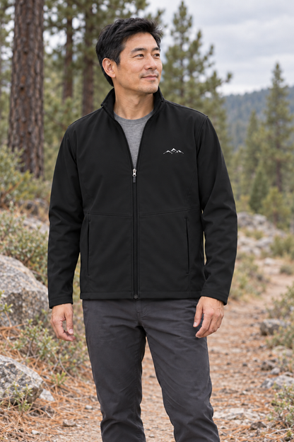 Illustrative lifestyle mockup: man wearing the Deep Black Men’s Mountain Softshell Jacket on a pine trail, showing its stand collar, full zip and small white mountain chest embroidery.