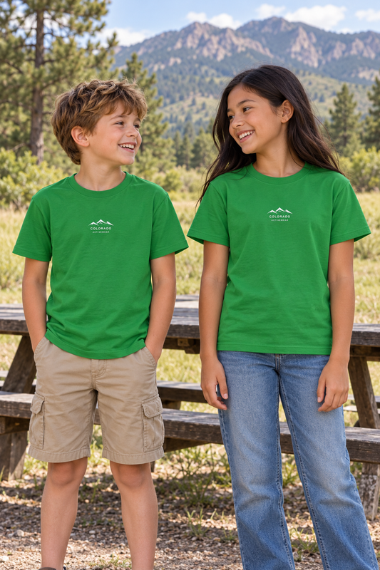 Illustrative lifestyle mockup: two school-age children wearing the Irish Green Colorado Mountain Youth Tee outdoors, showing its small white mountain chest logo.