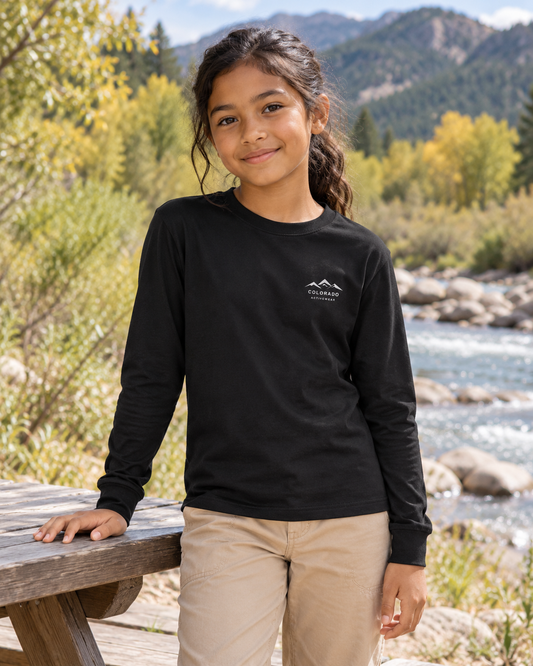 Illustrative lifestyle mockup: child wearing the Black Colorado Mountain Youth Long Sleeve beside a riverside picnic table, showing its small white mountain chest logo.